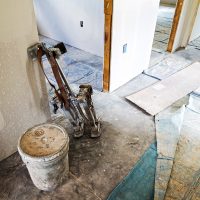 The drywall phase of a home renovation project.  Stilts and a bucket sit in a hallway with tarps partly covering the concrete floor.